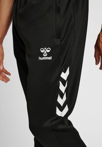 Svarta sportbyxor med en slät textur, utrustade med en vit logotyp och ett chevronmönster på sidan, designade för komfort och rörelse.