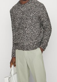 Jersey de punto con un patrón moteado en negro, blanco y tonos pastel. Presenta un cuello redondo y puños acanalados, combinado con pantalones verdes claros.