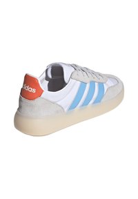 Vit sneaker med ljusgrå mockadetaljer och blåa ränder. Rund tå, snörningsdesign och orange hälflik med varumärkesnamnet.