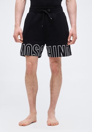 Homme portant un short noir à cordon avec un grand texte blanc "MOSCHINO" imprimé sur l'ourlet avant, debout pieds nus devant un fond blanc.