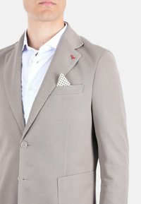 Blazer grigio chiaro realizzato in tessuto testurizzato, con due bottoni, tasca a patta e un fazzoletto bianco a pois. Accento rosso sulla bavetta.