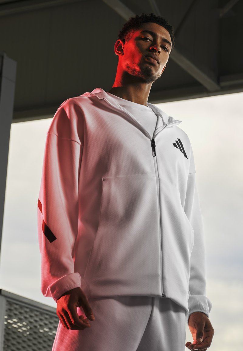 Veste à capuche blanche zippée avec une finition texturée, ornée d'accents de logo noirs et de reflets rouges. Associée à un pantalon blanc assorti.