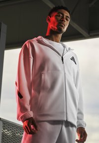 Młody mężczyzna w białej bluzie z zamkiem i spodniach, stojący pod konstrukcją, na której twarz i ubrania pada miękkie czerwone światło.