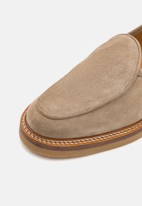 Béžová nubuková slip-on obuv s kulatou špičkou, dekorativním prošíváním podél podrážky a hladkým koženým podšívkou. Svršek obsahuje jemné logo.