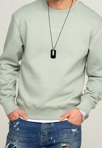 Hombre con sudadera verde claro que lleva el texto "Siempre Atemporal", jeans azules desgastados, reloj de pulsera plateado y collar de perro negro.