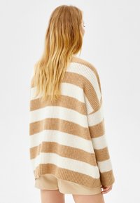 Kobieta z długimi blond włosami, ubraną w beżowy i biały sweter w paski oraz beżowe szorty, stojącą plecami do obiektywu na jednolitym tle.