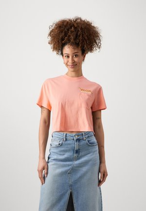 Pfirsichfarbener Crop-Top mit kurzen Ärmeln und einer Fronttasche, mit goldfarbenem "Volcom"-Logo. Kombiniert mit einem hochtaillierten blauen Jeansrock.