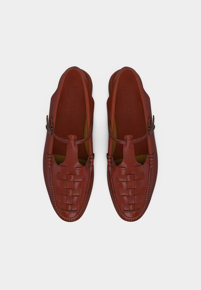 Καφέ δερμάτινα παπούτσια χωρίς κορδόνια με πλεκτή λεπτομέρεια μπροστά και T-φόρμα με πόρπη, εμφανιζόμενα από πάνω σε απλό φόντο.