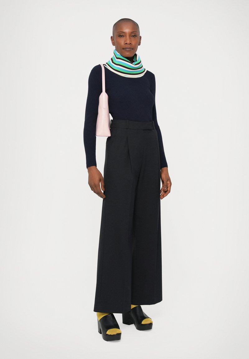 Marineblauwe geribde trui, meerkleurige gestreepte sjaal, zwarte wijde pantalons met plooien, gele sokken en zwarte plateau-schoenen. Roze handtas.