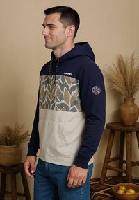 Hoodie met een navy bovenkant, een gepatterneerde beige en groene borstpaneel en een kangoeroezak. Inclusief een logo op de borst.