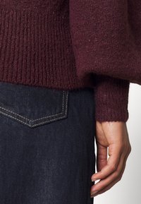 Viininpunainen neulepaita, jossa on resorikuvio, väljällä istuvuudella ja hihoissa käänteet. Yhdistetty tummansinisiin farkkuihin, joissa on takataskun yksityiskohta.