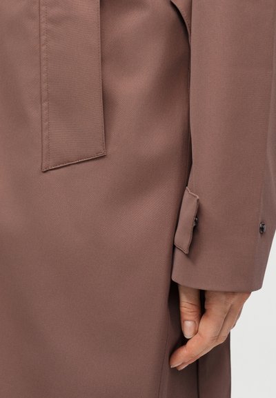 Κοντινό πλάνο σε μανίκι ταμπά παλτό με λεπτομέρεια ιμάντα με κουμπί και το χέρι ενός ατόμου που ακουμπά δίπλα στο παλτό.