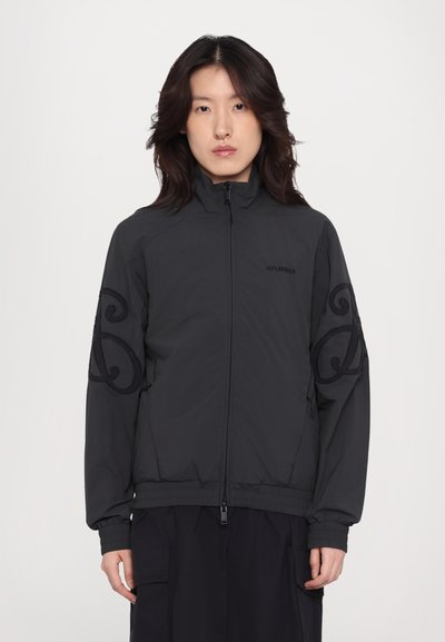 Zwarte zip-up jas van lichtgewicht materiaal. Heeft een kenmerkende oversized logo borduursel op de mouwen en ribgebreide manchetten.