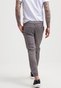 Man met getatoeëerde armen, gekleed in grijze joggingsbroek, zwarte sneakers en een wit T-shirt, die wegloopt tegen een effen witte achtergrond.