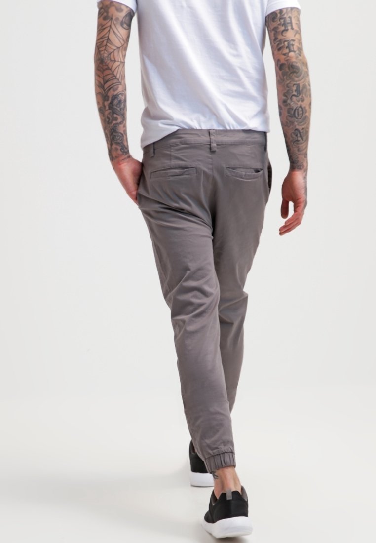 Man met getatoeëerde armen, gekleed in grijze joggingsbroek, zwarte sneakers en een wit T-shirt, die wegloopt tegen een effen witte achtergrond.