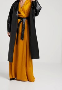 Black vinyl coat over a bright yellow silk dress, featuring a wrap design and matching belt, with a smooth texture and flowing silhouette.