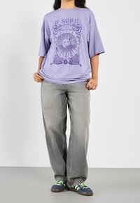 Camiseta gráfica lavanda con un diseño de sol, combinada con unos vaqueros grises holgados y zapatillas navy con rayas verdes. Ajuste casual y tela suave.