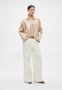 Béžový vlnený kabát s golierovým výstrihom, zapínaním na gombíky a prehnanými rukávmi, skombinovaný s krémovými širokými nohavicami a hnedými podpätkami.