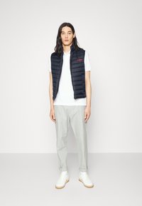 Chaleco acolchado azul marino con cremallera frontal, marcado en rojo, usado sobre una camiseta polo blanca, combinado con pantalones grises claros y zapatillas blancas.