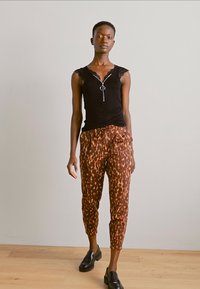 Black sleeveless top with ruffled edges, silver zipper detail; brown pants with abstract pattern; black shoes; wooden floor background.