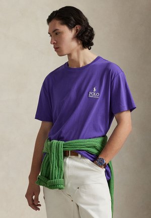 T-shirt en coton violet avec un col rond et des manches courtes, arborant un logo blanc brodé. Pull en maille vert noué autour de la taille.