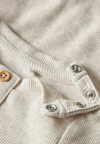 Geripptes, cremefarbenes Stoffmaterial mit Knopfverschluss und silbernen Druckknöpfen am Kragen. Weiche Textur mit dezenten horizontalen Streifen.