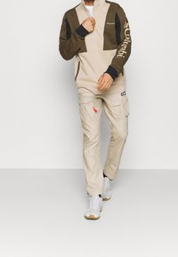 Giacca in fleece beige e marrone con zip a metà e dettagli neri, logo sulla manica; pantaloni cargo beige con dettagli delle tasche e etichetta rossa; sneakers bianche.