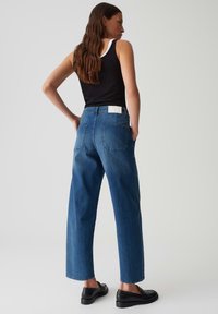 Hoge taille blauwe denim jeans met een rechte pijp, vijf zakken en een wit merkje aan de achterkant, gecombineerd met een zwarte tanktop.