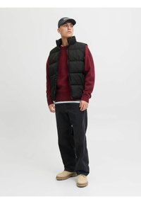 Chaleco negro acolchado sobre un suéter burdeos, combinado con pantalones oscuros y zapatos beige. Una gorra sencilla completa el conjunto. Fondo neutro.