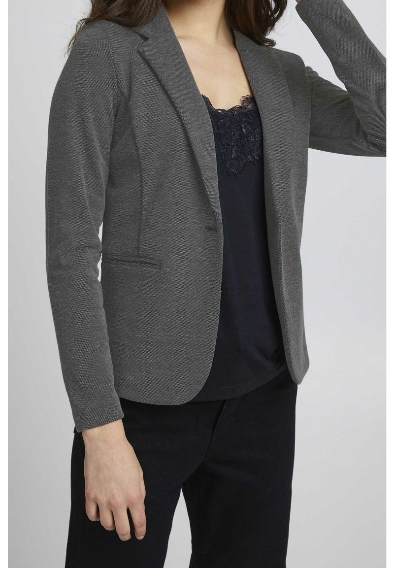 Femme portant un blazer gris sur un top noir orné de dentelle et un pantalon noir, un bras levé près de la tête.