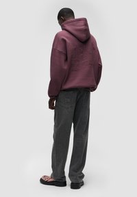Felpa con cappuccio di colore bordeaux con una grafica a trama sul retro, abbinata a jeans grigi a taglio largo con tasche ricamate.