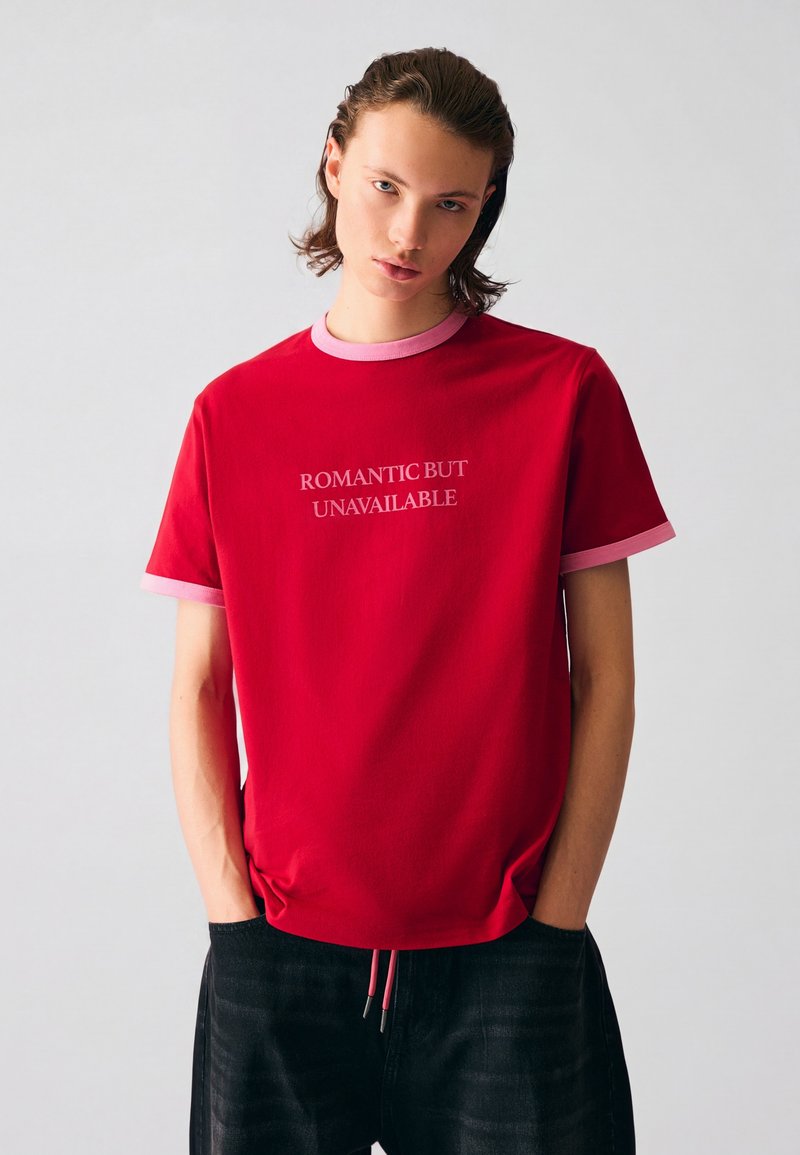 Jeune personne aux cheveux mi-longs portant un t-shirt rouge avec des bordures roses et le texte « ROMANTIC BUT UNAVAILABLE », debout les mains dans les poches.