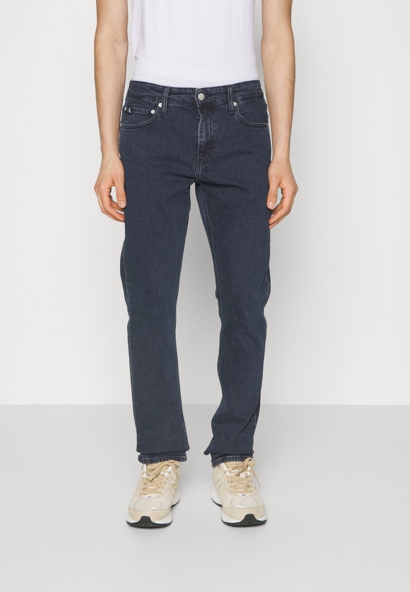 Mörkblå denimjeans med rak skärning, fem fickor och framknäppning, i kombination med ljusa träningsskor.