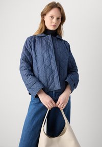 Blauwe gewatteerde jas met een brede kraag en twee knopen, gecombineerd met hoge taille blauwe spijkerbroeken. Houdt een crèmekleurige leren handtas vast.
