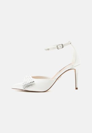 Hvid spids højhælet sko med ankelrem med spænde og dekorative sølvnitter på skoens forreste øverste del.