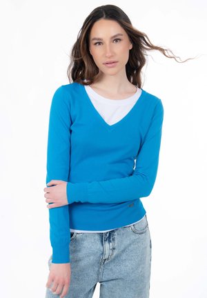 Suéter azul ajustado con escote en V y mangas largas, hecho de un material suave, superpuesto sobre una camisa blanca, combinado con jeans de mezclilla clara.