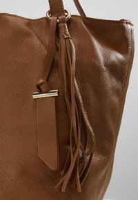 Sac à main en cuir brun texturé avec bandoulière longue, anneau doré et détail en pampille de cuir.