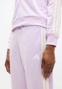 Αθλητική φόρμα σε λιλά χρώμα με παντελόνια στενής γραμμής και ταιριαστό μπουφάν. Περιλαμβάνει αντίθετες κρεμ ρίγες και ανάγλυφο λογότυπο.