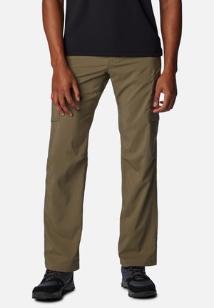 Mann trägt olivgrüne Cargohosen und schwarze Wanderschuhe, steht vor einem schlichten weißen Hintergrund.