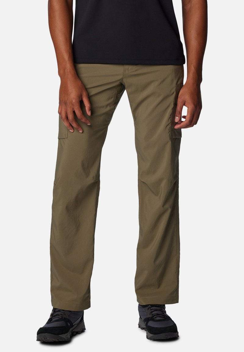 Uomo che indossa pantaloni cargo verde oliva e scarpe da trekking nere, in piedi davanti a uno sfondo bianco uniforme.