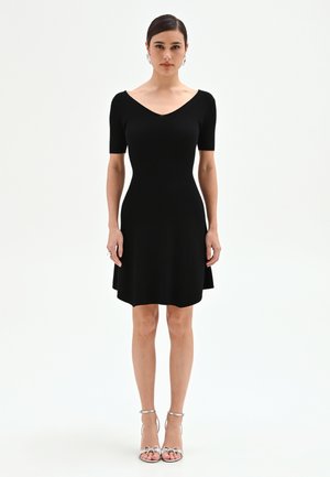 Femme debout, face à l'avant, portant une robe noire moulante à manches courtes et des sandales argentées à talons hauts sur fond blanc.