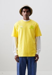 Camiseta de algodón amarilla con cuello redondo y mangas cortas, con detalles del logotipo en verde. Lleva puesta una camisa de manga larga blanca y jeans azules.