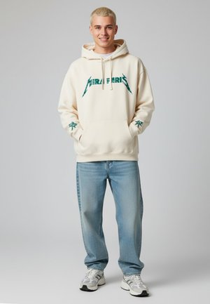 Crème-kleurige hoodie met groene grafische tekst, kangoeroezak, ribgebreide manchetten, gedragen met lichtblauwe jeans en grijs-witte sneakers.