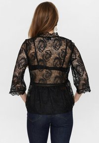 Blusa nera in pizzo con motivo floreale, maniche a tre quarti e orlo scalloppato. La parte posteriore è trasparente con stile peplum.