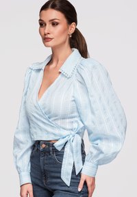 Femme aux cheveux bruns en queue de cheval portant un chemisier cache-cœur bleu clair avec des manches bouffantes et un jean foncé taille haute, tournée vers la gauche sur un fond uni.