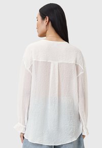 Hvid, letvægts bluse med en krøllet tekstur, lange ærmer, rund kant og detalje i ryggen ved sømmen. Lukket med knapmanchetter.