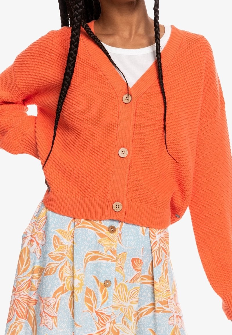 Oranger Strickcardigan mit V-Ausschnitt, versehen mit Holzknöpfen und einem strukturierten Design. Kombiniert mit einem Rock mit Blumenmuster.