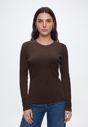 Vrouw met lang bruin haar draagt een donkerbruine longsleeve en blauwe spijkerbroek, staand tegen een effen lichte achtergrond.