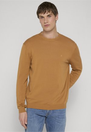 Giovane uomo con capelli corti castani che indossa un maglione senape a maniche lunghe e jeans azzurri, in piedi davanti a uno sfondo semplice.