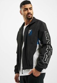 Black zip-up jacket with a white and blue panel. Features logo on chest, bold lettering on sleeve, and elastic cuffs and hem.